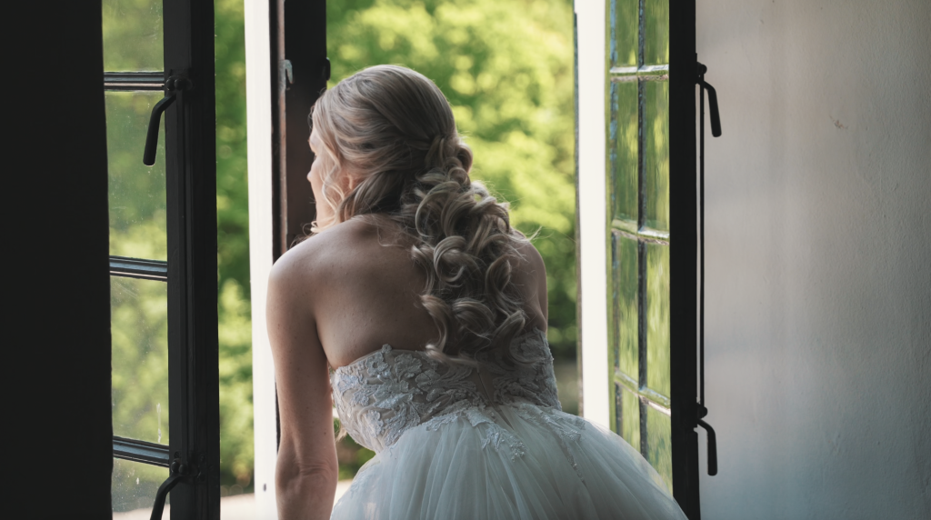 Bruid met een lange, golvende haarstijl, zittend bij een open raam in een lichte ruimte.