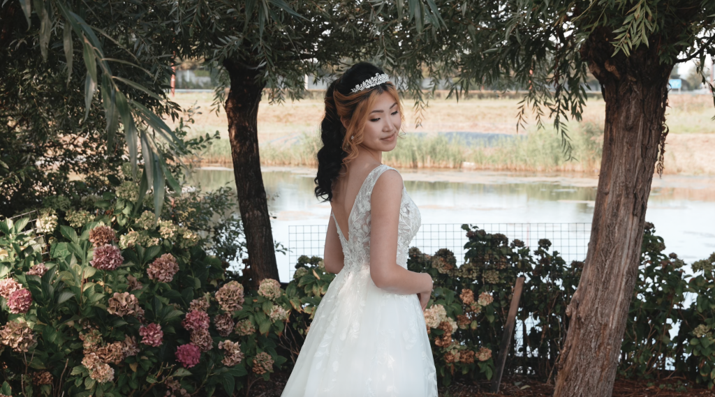 Aziatische bruid in een trouwjurk met een lage rug, staand bij een vijver met bloemen op de achtergrond.