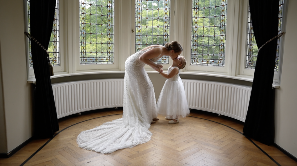 Bruid in een witte jurk kust een klein meisje in een witte jurk bij een raam met glas-in-lood.